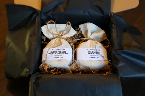 Two shave soaps, Palo Santo and Mystic Tobacco Vanilla, in muslin bags tied with jute string and labeled with white soap labels, inside a box lined with black tissue and brown crinkle paper