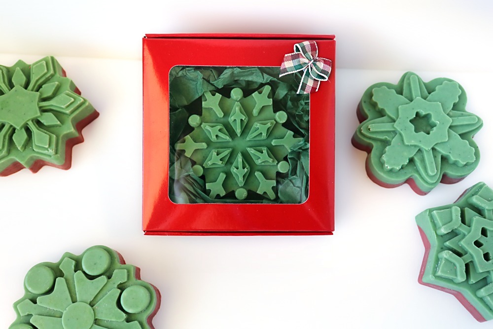 Green-topped snowflake soap in a red window gift box with green tissue paper and a plaid bow, surrounded by green-topped snowflake soaps