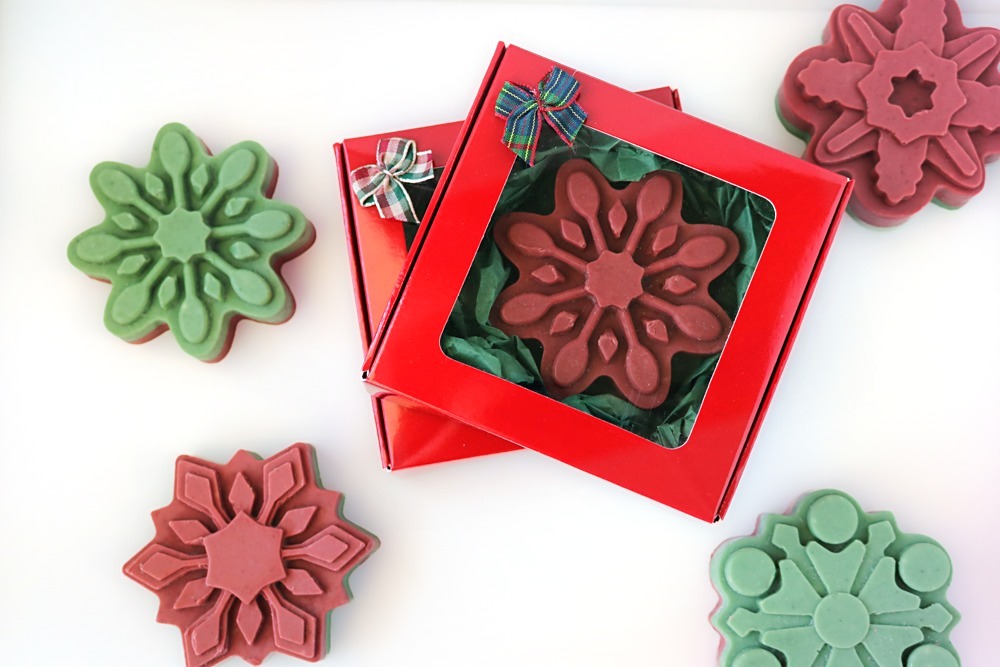 Close-up of a red and green layered snowflake soap with a red top, in front of a red gift box with more red snowflake soaps beside it