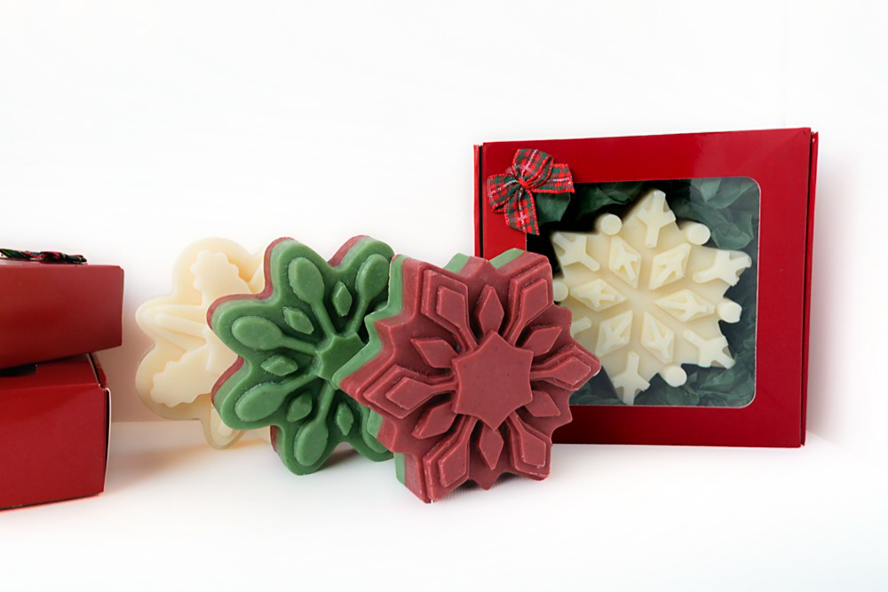 White, green top, and red top snowflake soaps together next to a red gift box with a plaid bow showing a white snowflake soap inside, with two stacked red gift boxes at the side