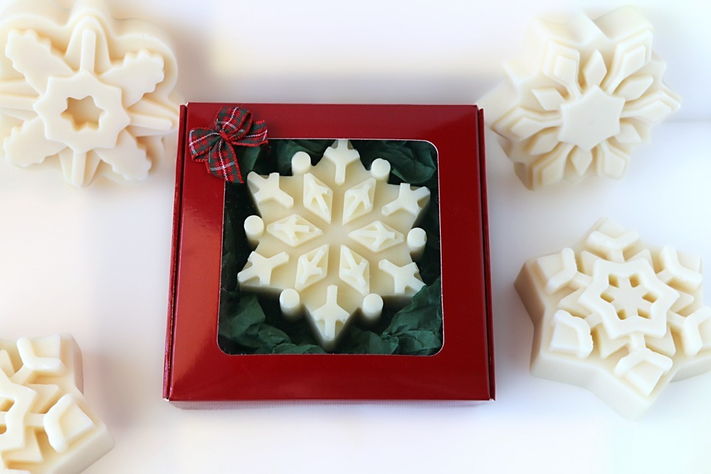 White snowflake soap gift in a red window box with green tissue paper and a small plaid bow, surrounded by white snowflake soaps in other designs