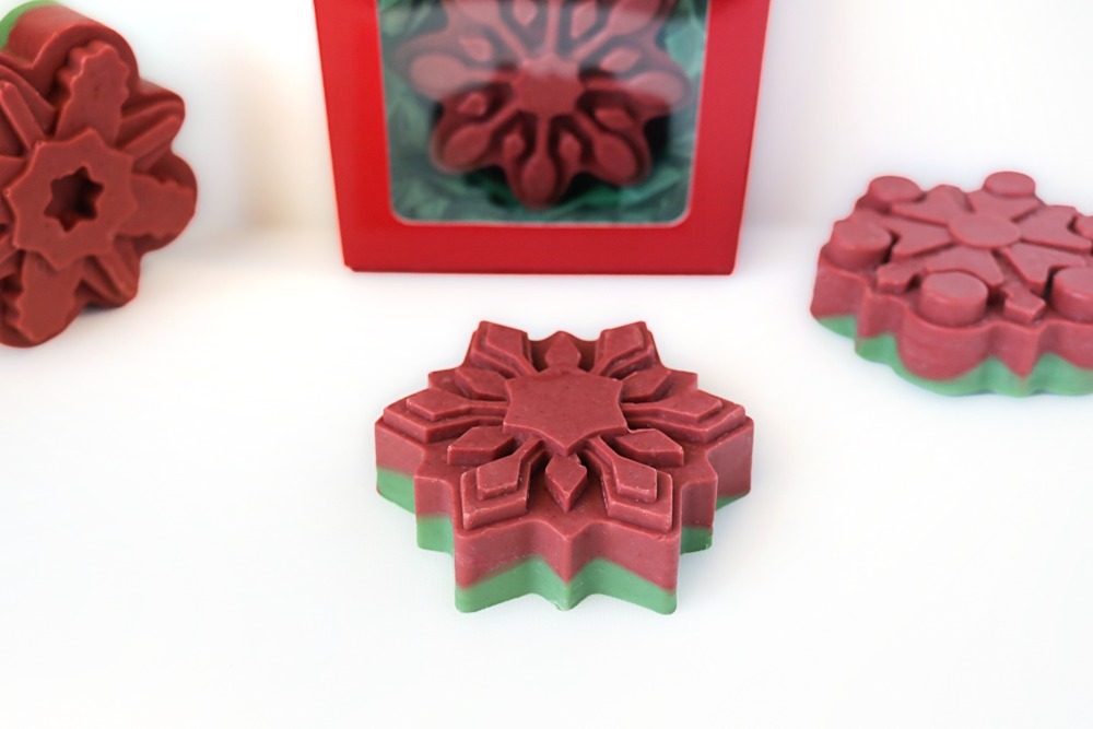 Close-up of a red and green layered snowflake soap with a red top and a green layer underneath, with more red snowflake soaps and a red gift box behind it